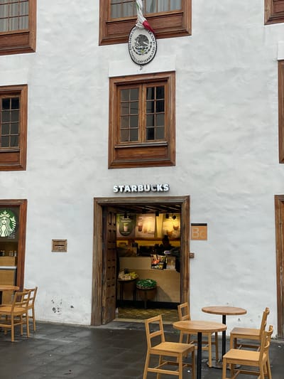 A street-level view captures a white-plastered building facade, featuring multiple rectangular windows framed in dark wood. Dominating the right side of the image is the entrance to a Starbucks coffee shop, marked by its vertical "STARBUCKS" sign in white letters on the left side of the wooden doorway. Above the entrance, a small orange sign with "Bc" is visible.
Through the open doorway, the interior of the Starbucks is partially visible, showing counters, menu boards, and various items. One poster inside appears to mention "SUAVE Y CREMOSA". Adjacent to the Starbucks entrance, an outdoor seating area is set up with several light-colored wooden chairs and two round, wooden-topped tables with dark bases. All chairs are empty, suggesting a quiet moment.
On the left side of the building's facade, above a wooden-framed window, hangs a circular emblem. Text clearly visible on the emblem reads "MEXICANOS UNIDOS" and "CONSULADO HONORARIO", along with an eagle crest, indicating the presence of a Mexican Honorary Consulate. Below the Starbucks sign, a small, recessed wooden box-like fixture is present on the white wall.
The ground in front of the building is paved with dark tiles. The overall scene blends traditional architecture with a modern commercial presence. The lighting suggests daytime, possibly overcast, as there are no harsh shadows.
