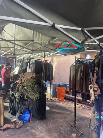 The image depicts an outdoor retail setup, possibly a market stall in Mira-Bhayander, India. Garments, appearing to be military-style clothing and accessories, hang on metal clothing racks under a white and gray pop-up tent. Below the hanging clothes, piles of folded dark-colored fabrics and olive drab gear are visible. A blue plastic bag sits on the ground, along with a pair of brown boots. A person wearing a red shirt and a baseball cap stands partially obscured near the bottom of the frame. An orange plastic basket is partially visible. The ground is a dark, rough surface. The light is diffused, suggesting an overcast day or a shaded area.
