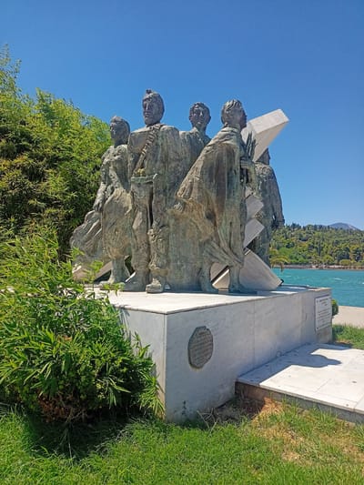 A bronze sculpture of five standing figures is positioned on a white marble pedestal. The figures are facing towards the right of the frame. The sculpture is located in Chalkida, Greece, near the sea. The sea is visible in the background, with trees and hills on the opposite shore. The sky is clear and blue. Green foliage and grass surround the base of the sculpture. There are two plaques on the marble pedestal, one circular and one rectangular. The rectangular plaque contains text. The overall scene is sunny and depicts an outdoor monument.