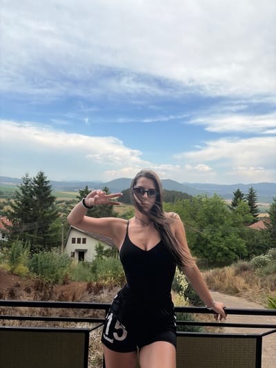 A young woman with long brown hair and sunglasses is posing for a photo on a balcony. She is wearing a black tank top and black shorts with "hummel 13" visible on the right leg. She is holding up two fingers in a peace sign with her right hand, and her left arm is resting on the balcony railing. The background shows a scenic view of rolling hills covered in trees and a small white house. The sky is partly cloudy with blue skies. The time of day appears to be daytime. The location is Vladislavtsi, Bulgaria. The woman appears happy and relaxed.