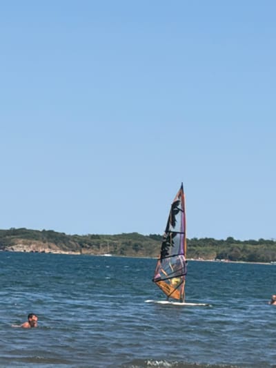 The image depicts a sunny day at a body of water, likely a bay or sea, with a distant tree-covered shoreline. In the foreground, a person is swimming in the blue water. To the right of the swimmer, a windsurfing board with a colorful sail is positioned on the water. In the mid-ground, another person is visible in the water to the right of the windsurf board. The sky is a clear, light blue. The scene is consistent with a recreational area along the coast of Atiya, Bulgaria, known for its beaches and water sports.