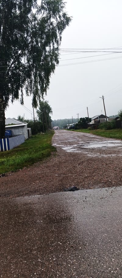 The image depicts a rural road in the village of Podgornoye, Russia, on a cloudy day. The road is unpaved and appears to be made of gravel, with several puddles of water indicating recent rain. The road curves to the right and leads into the distance, where more houses and trees are visible. On the left side of the road, there is a white building with a blue and white striped fence. A large tree with dark green leaves overhangs the left side of the image. Electrical poles and power lines are visible along the road, stretching into the distance. Houses with various roof types are scattered along both sides of the road. The overall scene suggests a quiet, possibly damp, rural environment.