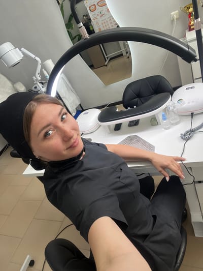 A woman with a black head covering, black scrub top, and black pants is sitting in a black chair at a white desk in what appears to be a nail salon. She is wearing a black face mask around her neck. A large circular lamp with a black arm is positioned above the desk, illuminating the scene. On the desk are several items related to nail care, including a white UV lamp with a digital display, a hand rest, a bottle of clear liquid, and a mesh dust collector. A large mirror is visible in the background, reflecting the room. The flooring appears to be light-colored tiles. There is no visible indication of the city of Stepnogorsk or its administration in the image.