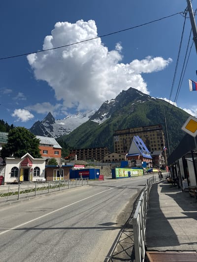 This image depicts a street scene in Dombay, Russia, with a dramatic mountain backdrop. The road runs alongside a row of buildings, some of which appear to be shops or businesses. One building has a distinctive blue roof. There are also shipping containers visible, suggesting commercial activity.

In the foreground, a metal fence lines the road. On the left side of the road, a large, snow-capped mountain dominates the sky, its slopes covered in green vegetation. Several white clouds are scattered across the clear blue sky. Power lines are strung overhead.

A flag, possibly the Russian flag, is visible in the upper left corner of the image. Signs on the buildings are in Russian. One sign reads "СТОЛОВАЯ," meaning "canteen" or "cafeteria," and another appears to be "МАГАЗИН," meaning "shop." A sign with a yellow diamond shape, often used for warning signs, is also present.

The time of day appears to be daytime, with bright sunlight casting long shadows. The weather seems pleasant, with clear skies and sunshine. The overall impression is a bustling mountain resort town. There are no people prominently featured in the image.