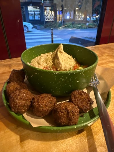 A close-up, eye-level shot shows a green bowl filled with hummus and chopped vegetables, accompanied by several falafel balls on a matching green plate. The food is placed on a wooden table, with a blurred background showing a street scene through a window, including a crosswalk, a car, and storefronts. The image is taken indoors, likely in a restaurant, in the town of Bellingen, Australia. The overall lighting suggests it is evening or dusk.