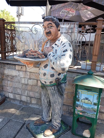 A full shot depicts a statue of a man holding a platter of food, located in the city of Chinarkent, Uzbekistan. The statue is positioned on a stone-tiled ground, and a brick wall serves as the background. To the right of the statue, there is a green metal box with a religious advertisement. The statue is of a man with a mustache, wearing a patterned shirt and gray pants, and a hat. He holds a platter of what appears to be cooked meat or sausages. In the background, an outdoor seating area with chairs, tables, and an umbrella with "PEPSI" on it are visible. In the background, there are people on the seating area. A wrought-iron fence is adjacent to the statue on the left.