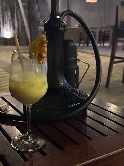 A sideways view showcases a frosty yellow cocktail garnished with a pineapple wedge and straw, sitting on a wooden slatted surface. Behind the drink, a black metal structure with a looped handle and cylindrical component dominates the frame. The backdrop is blurred, suggesting an outdoor setting with faint hints of architectural elements and vegetation. The scene is lit softly, casting a cool hue.