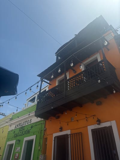 The image shows the exterior of two colorful buildings on a bright, sunny day in Old San Juan, Puerto Rico. The building on the left is painted bright green with white trim around two doorways. Above the doorways, signage reads "ANTOJITOS DEL CALLEJON" in white letters. The building to the right is painted a vibrant orange. A dark wooden balcony with intricate latticework spans across the upper floor of both buildings, supported by dark wooden beams. String lights with bare bulbs are draped across the balcony and extend across the foreground. In the distance, under a clear blue sky, a black umbrella is partially visible on the left. The scene depicts a street or alleyway with commercial establishments, likely restaurants or shops, characteristic of the historic district.