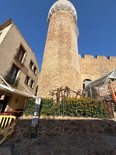 The image shows a close-up, low-angle view of the Torre dels Moros, a medieval watchtower in Tossa de Mar, Spain. The tower, constructed of rough-hewn stones, is partially covered with a white mesh netting, suggesting ongoing renovation or preservation work. Adjacent to the tower is a tan-colored building with multiple windows and balconies. In the foreground, a metal fence encloses a garden with vibrant orange flowers. A metal sculpture, possibly an anchor, rests on the ground behind the fence. A informational display board is also visible in front of the garden. Scaffolding and plastic sheeting are present, indicating construction or maintenance activities on a stone wall structure to the right of the tower. The sky is clear and blue.