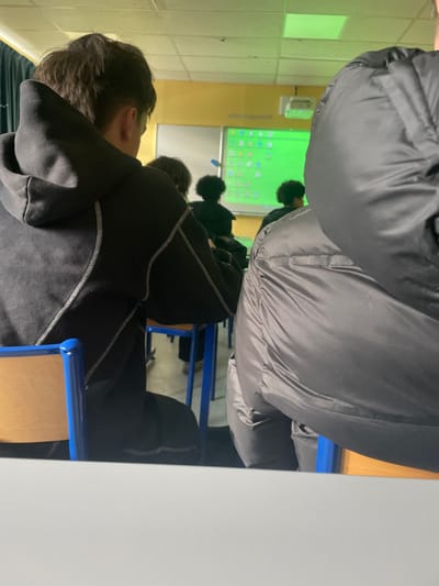 The scene depicts the interior of a classroom in Hérouville-Saint-Clair, France, viewed from a seated position. In the immediate foreground, a light grey table surface is present. To the left, the back of a person is visible, wearing a dark grey or black hooded sweatshirt with distinctive light grey double-stitched seams that form a V-shape across the upper back and extend along the sleeves. This individual has dark brown, medium-length hair and is seated in a chair with a light brown wooden backrest and a blue metal frame. To the right, the partial back and shoulder of another seated individual wearing a dark grey or black puffer jacket are visible.
Further into the room, several other individuals are seated at desks, facing the front. Their backs and heads are visible, with at least two individuals exhibiting dark, possibly curly, hair.
The front of the classroom features a large white surface serving as a projection screen. It displays a bright green computer desktop background populated with numerous application icons. A white overhead projector is mounted on the white ceiling, projecting onto this screen. The wall above the projection screen is painted yellow. Rectangular fluorescent light panels are integrated into the white ceiling, with some casting a greenish tint, likely from the projector's light spill. A dark fabric or wall section is visible on the far left of the frame. All visible individuals are seated and oriented towards the front of the classroom, indicating their attendance at a lesson or presentation.