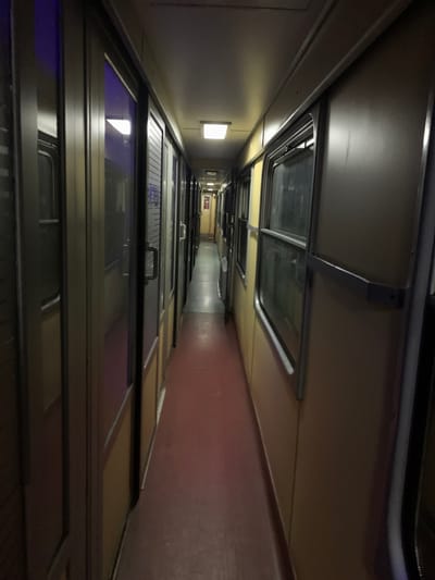 A dimly lit corridor stretches into the distance, lined with cabin doors on one side and a plain wall on the other. The doors have reflective windows and handles. A red-toned floor runs along the length of the corridor. Ceiling lights are sparsely placed, creating a sense of depth as the corridor narrows towards the end. The perspective emphasizes the length and linearity of the space.