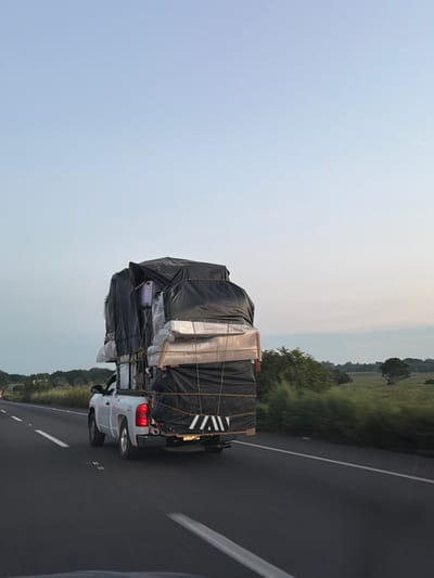 A white pickup truck overloaded with furniture and belongings is seen driving on a highway. The cargo is stacked high and covered with a black tarp, secured by ropes. The highway is adjacent to a field and sky. The truck is facing away from the viewpoint.