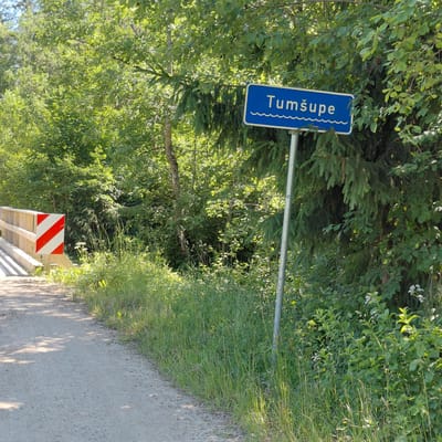The image shows a road in Skuķīši, Latvia, with a blue sign that reads "Tumšupe" with wave symbols below the text. The sign is mounted on a metal pole. To the left of the sign, a wooden bridge is visible with a red and white striped marker on its railing, indicating a hazard. The road is unpaved and appears to be a gravel path. Lush green vegetation, including trees, bushes, and tall grass, lines the sides of the road. Sunlight filters through the trees, casting shadows on the path. The environment is natural and appears to be in a rural or forested area. There are no people or animals visible in the image.