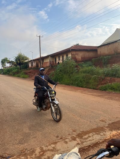 A person is riding a motorcycle on a paved road. The rider is wearing a black jacket, black pants, a blue cap with white lettering, and light-colored shoes. The motorcycle appears to be older, with a visible engine and exhaust pipe. In the background, there is a weathered building with a rusted metal roof, situated on a reddish-brown slope covered in dense green vegetation. Overhead, power lines stretch across the frame, supported by wooden poles. The sky is partly cloudy, suggesting it could be daytime, possibly morning or afternoon. The scene appears to be in Gembu, Nigeria, indicated by the presence of the building style and vegetation common in the region. The road is a light brown asphalt with visible gravel. The overall atmosphere suggests an everyday activity in a semi-urban or rural environment.