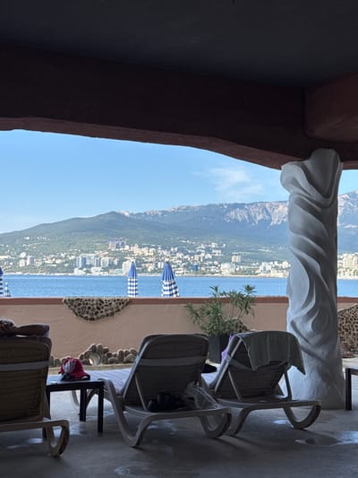 The image displays a sheltered outdoor terrace, located in Почтовское сельское поселение, Ukraine, offering a panoramic view of a large body of water (likely the Black Sea), a coastal city, and a distant mountain range.
The foreground depicts a concrete or tiled floor with visible wet patches. Three light-colored sun loungers are present; one on the far left is partially occupied by a person resting, clad in dark clothing. Adjacent to the central lounger, a small black side table holds a red hat and a dark object. The rightmost lounger has a light-colored towel draped over its backrest, and dark objects, possibly shoes, are visible beneath it. A potted plant with green foliage is situated between the sun loungers and a decorative low wall featuring embedded pebbles in a wavy pattern. A prominent white, ornate, twisted column stands on the right side of the frame, supporting a dark, flat overhead structure that frames the upper portion of the view.
Beyond the terrace, several blue and white striped beach umbrellas are visible on or near the water's edge, some partially closed. A dense urban area with numerous multi-story buildings extends along the coastline. Behind the cityscape, a large range of rugged mountains rises under a clear blue sky with minimal cloud cover. The scene is illuminated by bright daylight, with the terrace area appearing to be in shadow.