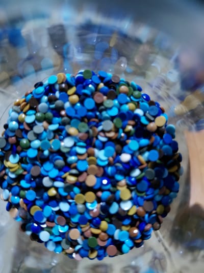 The image shows a close-up, top-down view of a pile of small, colorful, circular beads inside a transparent container. The beads are predominantly shades of blue, with some brown, yellow, green, and gray ones mixed in. The background is blurred and appears to be a reflective surface, possibly glass or plastic, with light creating bokeh effects. The scene is not identifiable as being in Ертіс ауылдық округі, Kazakhstan, as there are no specific landmarks or contextual clues to indicate this location. The image is purely a still life of beads.