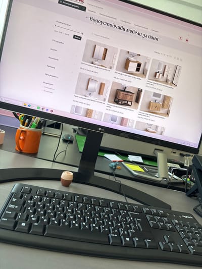 This media file captures a close-up view of a computer monitor displaying a website for bathroom furniture. The screen shows various bathroom cabinets and vanities with their prices and descriptions in Bulgarian. The website also includes filter options for price, product type, design, availability, and manufacturer.

In the foreground, a black Logitech keyboard is prominently featured, angled towards the right side of the frame. To the right of the keyboard, a small, light-brown, round object rests on the desk. The background is dominated by the computer monitor, which is displaying the e-commerce website. A black computer mouse is visible on the desk behind the monitor. In the lower right corner, an orange ceramic mug filled with pens and markers is positioned.

The overall scene suggests a home office or workspace environment. The lighting appears to be artificial, typical of an indoor setting. The timestamp on the monitor indicates "11:06" and "20.6.2026 г.", suggesting it is daytime on June 20, 2026. There are no people visible in the image. The primary focus is on the computer setup and the online shopping interface. The visible text is primarily in Bulgarian, related to furniture products and website navigation. The location context of Busmantsi, Bulgaria, provides a geographical setting for this scene.