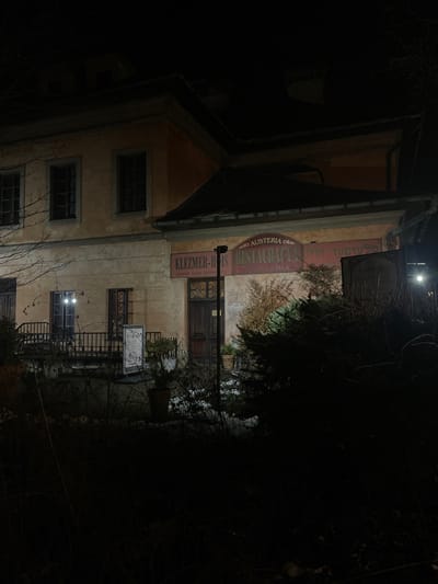 The image displays an old, two-story building at night, seen from a slightly upward angle. The building has a light-colored, possibly beige or pale yellow, facade with a dark, sloped roof or awning extending over the left side. Several windows with dark frames are visible on both floors.
A large, reddish sign with white lettering is prominent on the upper part of the facade. The text clearly reads "RESTAURACJA," with "AUSTERIA" written in smaller letters above it. Below this main sign, a vertical red sign spells out "KLEZMER-HUIS," followed by smaller, legible text that says "MUZYKA ŻYDA." A dark wooden entrance door is centrally located on the ground floor. To the right of the entrance, a decorative metal railing extends, and a freestanding sign or menu board, with illegible text, stands further to the right.
The scene is dimly illuminated by artificial lights, casting shadows and highlighting parts of the building, confirming it is nighttime. Dark, dense trees and branches partially obscure the right side and foreground of the building, contributing to the nocturnal atmosphere. Some sparser, lighter vegetation is visible near the entrance. The building's architecture and the text on the signs ("AUSTERIA RESTAURACJA KLEZMER-HUIS MUZYKA ŻYDA") indicate a historic restaurant or inn with a Klezmer music theme, located in Kraków, Poland. The overall impression is quiet and still.