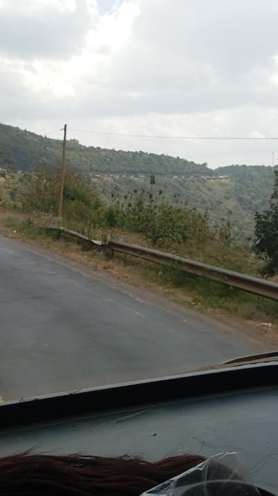 The scene depicts a view from within a vehicle looking towards a landscape in Lari/Kirenya ward, Kenya. In the immediate foreground, strands of dark reddish-brown hair and a crumpled transparent plastic bag are visible on a dark, flat surface, consistent with a vehicle's dashboard. A two-lane asphalt road, appearing damp, curves gently uphill and to the left, occupying the lower-mid portion of the image. Faint white line markings are discernible on the road's right edge. Parallel to the road on the right, a rusty metal guardrail is present, appearing partially detached from its post near the foreground. The roadside terrain is unpaved, covered with dry grass and sparse green shrubs. A wooden utility pole stands on the right, alongside the road, with thin wires extending horizontally across the mid-ground. Beyond the road and immediate roadside, a hilly landscape rises, densely covered with green and brownish vegetation, including numerous shrubs and small trees. Along a distant ridge or road on the hillside, a linear arrangement of numerous small, light-colored structures, consistent with residential dwellings, is visible. The sky is largely obscured by a dense layer of gray and white clouds, indicating overcast conditions. No individuals are directly visible.