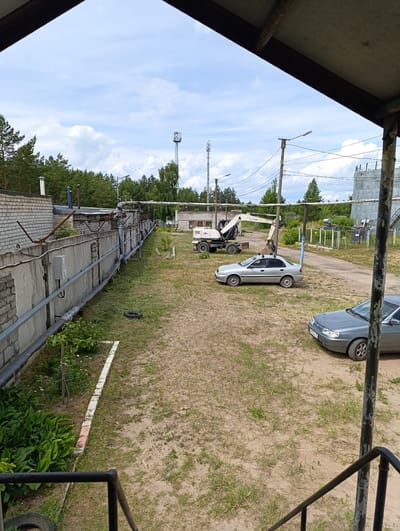 The image shows an outdoor industrial area with a view from an elevated vantage point, possibly a balcony or walkway, looking down on a dirt and grass lot. In the foreground, metal railings are visible. The scene includes a long, low concrete wall running along the left side, with pipes mounted on it. Beyond the wall are some buildings constructed of brick and concrete.
In the middle ground, a white excavator with the brand "JUM" is parked on the dirt. Next to the excavator is a light silver sedan. Further into the scene, a darker colored sedan is parked on the right side of the lot. In the background, there are trees, a large concrete cylindrical structure, and utility poles with wires and antennas. The sky is partly cloudy. This appears to be an industrial or commercial property located in the city of Volzhsk, Russia, likely in an area with a mix of industrial facilities and natural vegetation. There are no people visible in the image.