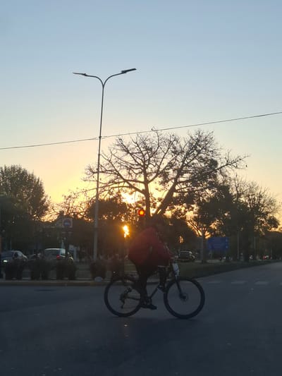 A street scene during sunset in Villa Domínico, Argentina, shows a person riding a bicycle on the road. A silhouetted tree is positioned between the setting sun and the camera. A street light is visible on the left. Cars and pedestrians are present on the right side of the road near a street sign. The sky is gradient with light blue and orange hues.