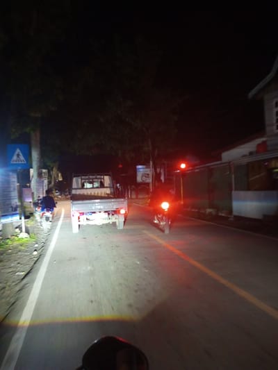 A low-angle, night-time, first-person perspective shot captures a street scene in Manurungnge, Indonesia. The foreground is dominated by the handlebars and a portion of the seat of a motorcycle. In the mid-ground, a white pickup truck is traveling away from the viewer, its taillights illuminated. To the left of the truck, a pedestrian crossing sign with a blue background and a white pedestrian symbol is visible on a pole. Further down the road, another motorcycle, with its headlight on, is moving towards the viewer. The street is asphalt, with a white line marking the lane. Buildings with lit windows are visible on the right side of the road. Trees line the left side of the road, casting shadows in the darkness. The scene is illuminated by the headlights of vehicles and streetlights, creating a reddish glow on the road surface.