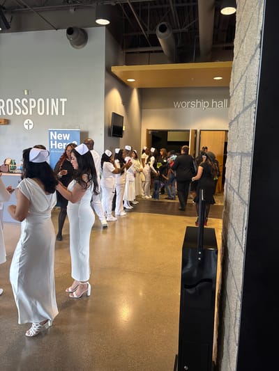 This media file captures an indoor gathering in Grand Prairie, United States of America, likely within a church or community center. The scene is dominated by a group of individuals, predominantly women, dressed in white attire, including dresses, pantsuits, and scrubs, many wearing white caps resembling traditional nurse caps. They are arranged in lines, suggesting an organized event or ceremony, possibly a graduation or induction for healthcare professionals, given their uniform appearance.

In the foreground, two women in white dresses and caps are visible, one adjusting her attire. Further back, more individuals in similar white outfits are lined up. In the background, other people are seen in casual, darker clothing, observing the event or waiting.

The setting is a modern indoor space with light-colored walls (grey and yellow/beige) and a polished concrete or light brown floor. The ceiling features exposed industrial-style structures and circular light fixtures. Visible on the wall is a large sign with the text "CROSSPOINT" and a cross symbol, indicating a church. Another banner features the text "New Heroes" and "We're here to". In the background, a sign reads "worship hall," confirming the nature of the venue. A black podium or stand is also present. The lighting is bright and artificial, suggesting an indoor event at an indeterminate time of day. The atmosphere appears formal and celebratory.