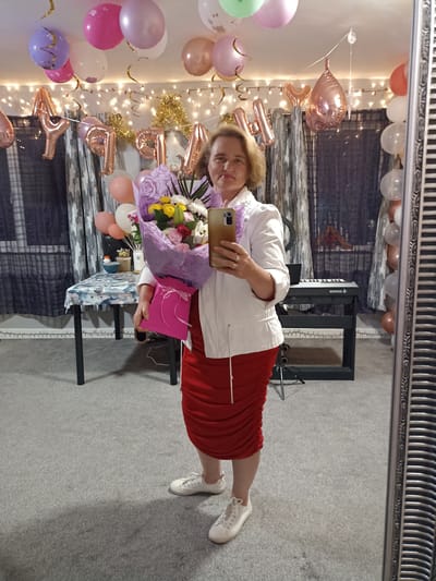 In the image, a woman stands in what appears to be a domestic setting, possibly a living room or party space, decorated with balloons and fairy lights. She is holding a bouquet of flowers and a pink gift bag. The woman is wearing a white jacket, a red ruched skirt, and white sneakers. In the background, a table is set with various items, and a keyboard is visible. A large ornate mirror reflects the scene, showing more balloons and the woman from a slightly different angle. The overall ambiance suggests a celebration, potentially a birthday or anniversary. The specific location in Annagry, Ireland, is not determinable from the visual information provided.