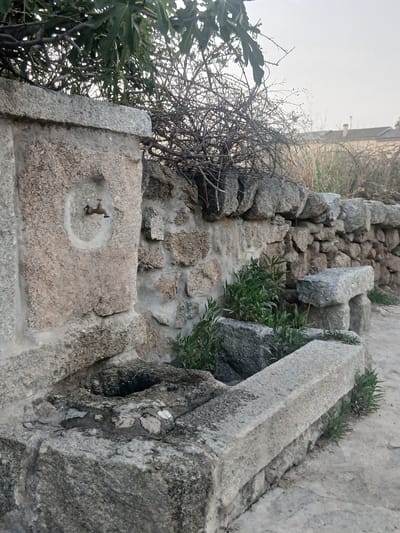 An old stone fountain is situated outdoors. The fountain is made of rough-hewn stone blocks, with a spout on the left side and a basin in the foreground. A wall constructed from similar stones forms the backdrop. Green vegetation grows around the fountain and along the wall. In the distance, buildings with roofs and chimneys are visible. The sky is overcast. The image depicts an aged, rustic water source in a natural setting, likely in the town of Navaluenga, Spain, given the provided context of its location.