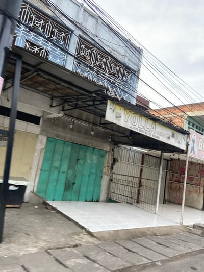 The image shows the exterior of a closed storefront located on Jl. Kopo Sayati No. 187 in Kecamatan Margahayu, Indonesia. The store is called "Yolla Setial", and is partially obscured by an awning. The building has a concrete facade and a teal-colored door.  The store is currently closed. There is a small white tiled area in front of the building. The environment is characterized by numerous power lines and a cloudy sky. There are no people visible in the image.  The ground is made of gray concrete and there are a few small, loose pieces of debris in the area in front of the store.  The image is slightly blurred, but the building and signage are clearly visible.