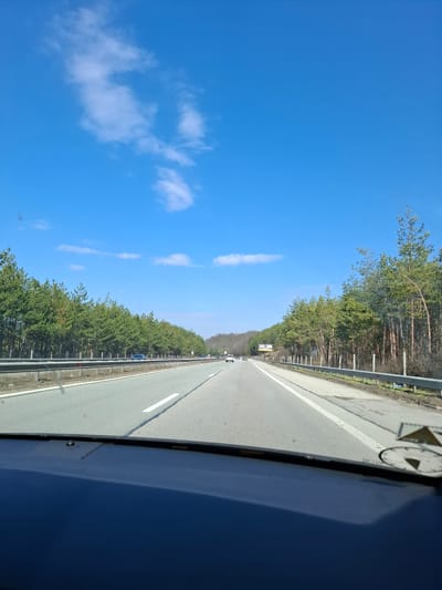 The image shows a view from inside a vehicle moving along a highway. The highway has two lanes and is bordered by trees and a metal guardrail. The sky is blue with some clouds. There are other vehicles visible in the distance on the highway. The view is from the passenger side of the vehicle. The location is inferred to be somewhere near Gorna Vasilitsa, Bulgaria, based on the prompt, although no details within the image directly confirm this. No people are clearly visible in the image.