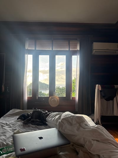 An indoor scene in Koh Tao, Thailand, features a room with dark wooden paneling on the walls and ceiling. In the immediate foreground, a disheveled bed with white and grey bedding is present. A silver Apple MacBook laptop, open and resting on a small stand, occupies the lower-left portion of the bed, accompanied by a green packet. A dark garment is crumpled on the bed's center.
A large, wooden-framed window dominates the midground, with bright sunlight streaming through its multiple vertical panes. A straw hat hangs centrally on the lower window frame. Translucent grey curtains are drawn to either side of the window. The exterior view reveals a tropical landscape with a prominent green, conical island rising from the sea under a partly cloudy sky. Lush green trees are visible closer to the window.
To the right, a white Mitsubishi air conditioning unit is mounted high on the wall. Below it, a clothes drying rack holds various garments, including white towels and dark clothing items. A partial view of a dark furnishing and a small table with bottles is visible on the far left. The scene is illuminated by strong natural light originating from the window.