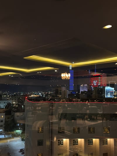 The image depicts a night view from a high vantage point within a city, likely Amman, Jordan, as suggested by the architectural style and building lights. The scene is characterized by a warm, inviting interior reflected in a large glass pane, juxtaposed with the cooler, distant city lights.
The foreground is dominated by a reflective surface, likely a window or glass partition, which displays a mirrored image of an indoor setting. This interior features a dimly lit space with what appears to be seating arrangements, possibly tables and chairs, suggesting a restaurant or lounge area. A prominent chandelier with multiple lights is reflected, adding a touch of elegance. The ceiling of this interior space is adorned with recessed yellow lighting, creating a warm glow.
Beyond the reflective surface, the city unfolds under the night sky. Numerous buildings of varying heights are visible, their windows lit with a mixture of white and yellow lights, indicating urban activity. Tall buildings with distinct architectural features, including one with vertical blue lights and another with horizontal red lights, stand out against the cityscape. The distant horizon is dotted with more lights, suggesting a sprawling urban environment.
A red light strip runs along the top edge of a building in the mid-ground, possibly an accent feature. Below this, the exterior of a multi-story building is partially visible, with windows that have an interesting geometric pattern. In the lower left portion of the image, a street or parking area is illuminated, with at least two cars visible. Vegetation is also present, adding a touch of green to the urban landscape. The overall impression is a vibrant city at night, viewed from a comfortable and stylish indoor setting.