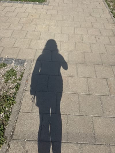 The image shows a shadow of a person cast on a paved walkway. The walkway is made of rectangular concrete pavers laid in a staggered pattern. There is a strip of gravel and some sparse green vegetation along the edge of the walkway. The shadow depicts a person standing, with their head and torso clearly visible, as well as outstretched arms or hands. The lighting suggests it is daytime, likely sunny, as indicated by the sharp shadow. The scene is an outdoor setting, possibly a park, sidewalk, or public square. The overall impression is that of a casual moment captured in sunlight. No text is visible in the image.