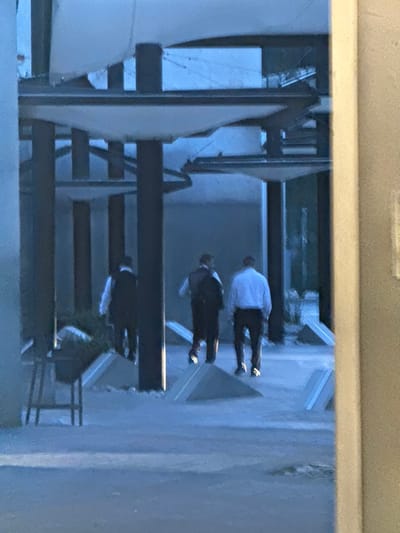 The image displays three adult males walking away from the camera through a structured outdoor or semi-outdoor space in Santa Catarina, Mexico. The scene is characterized by dim, bluish ambient lighting. The individuals are dressed in light-colored button-up shirts and dark trousers; the central figure wears a dark backpack, and the leftmost figure wears a dark vest. They are traversing a light-colored paved pathway. The pathway is bordered by multiple low, angled, triangular concrete barriers or planters, which contain light-colored gravel. Overhead, a modern architectural canopy structure is visible, composed of dark vertical support poles and horizontal beams, supporting several irregularly shaped, white fabric panels. A dark, three-legged stand is positioned to the left of the pathway. Distant vegetation is faintly visible in the background. No observable interactions between the individuals are depicted.