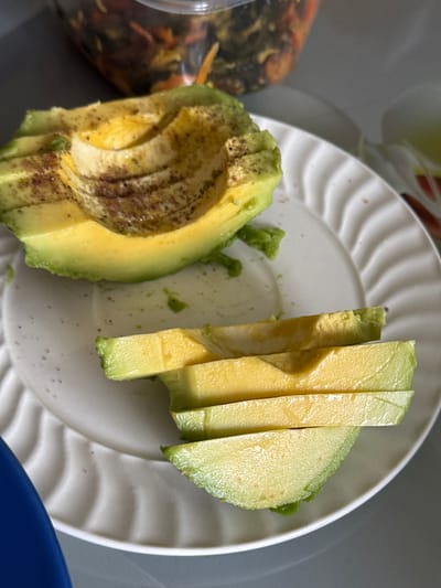 A close-up, overhead shot features a sliced avocado served on a white ceramic plate with a decorative wavy rim. The avocado is divided into two main parts: on the left, one half of an avocado, with its seed removed, lies flat on the plate, showing its pale yellow-green flesh dusted with a dark brown, speckled seasoning. On the right side of the plate, several long, thin slices of avocado are neatly arranged, standing upright, showcasing their vibrant green skin and creamy interior.
The white plate itself shows some scattered small brown specks, likely residue from the seasoning applied to the avocado. In the blurred background to the left, a translucent plastic container with a mix of dark and lighter-colored food items, possibly pickled vegetables or a salad, is partially visible. A hint of a blue object, likely another dish or bowl, is visible in the bottom right corner of the frame. The overall lighting suggests an indoor setting. No people or visible text are present in the image.