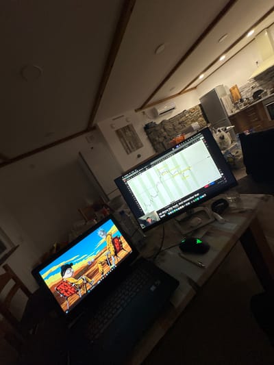The image depicts an indoor workspace with two monitors, a laptop, and a computer mouse on a desk. One monitor displays what appears to be a financial chart, while the other displays an animated cartoon. The desk is cluttered with miscellaneous objects, including a pen and some discarded items. The location is suggested to be Kisyovtsi, Bulgaria, though there is no direct evidence to support this claim from the image itself. The environment is dimly lit, creating a contrast between the bright screens and the surrounding darkness. There are no visible people in the image, and the focus is primarily on the technology and the workspace setup.