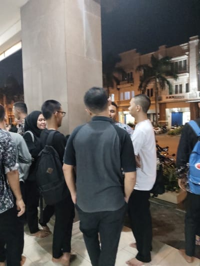 The image shows a group of people standing in front of a building in Telukjambe, Indonesia. The scene is lit by artificial lighting, with a nighttime setting.  The group is standing in front of a building, likely an entrance. The building is light brown with an off-white trim, possibly stucco or concrete. The building is two stories and has several windows and a door. The people appear to be casually conversing, their faces are not visible. They are standing in front of a glass door that leads to a dimly lit hallway with a tiled floor. There are six men, one woman wearing a hijab, and one man in the background wearing a blue backpack. One of the men is wearing a black backpack and a grey T-shirt, while the other man is wearing a white T-shirt. The people appear to be in their late teens or early twenties, and they are all casually dressed. They are standing on a tiled surface.  There is an unknown, possibly metal, object, to the left of the group in the bottom of the image, but it is obscured by a person’s leg.  A sign is visible over the building, but the text is too blurry to decipher. The scene has a sense of normalcy and is likely in the evening. The surrounding environment is not readily visible, but it appears to be a built-up urban area with a dark road and trees. The scene appears to be from a place in Telukjambe, Indonesia, at the coordinates -6.308, 107.282.