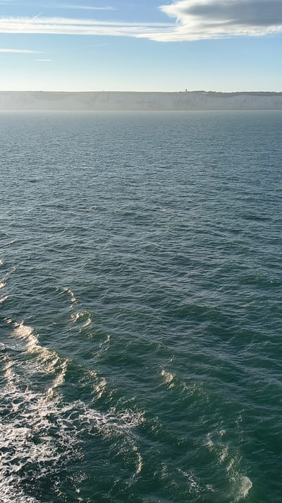 This is a photograph of the sea, with a distant view of the White Cliffs of Dover. The sky is blue with some clouds, and the sun is shining, creating reflections on the water. In the foreground, there are white waves and seafoam, indicating movement. The White Cliffs are visible in the distance as a pale line across the horizon. There is a small structure, possibly a lighthouse or building, visible on top of the cliffs. The sea is a deep blue-green color, with ripples and waves across its surface.
