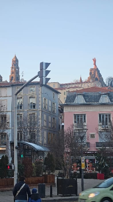 This image depicts an urban scene in Le Puy-en-Velay, France, with prominent landmarks in the background. The perspective is from a street level, looking towards higher ground.
In the foreground, two pedestrians, an adult and a child, are seen from behind, walking towards the right side of the frame. The adult wears a dark jacket and a white cross-body bag strap. The child wears a blue jacket and a blue hat, holding the adult's hand. To their right, the front portion of a light green compact car, blurred by motion, travels right-to-left. A black traffic light pole is situated in the lower left, displaying a green light for vehicles and a red "standing person" symbol for pedestrians. Several large planters containing bare trees and small shrubs are arranged along the street. Some planters are rectangular and made of rusty metal, while others are dark square pots.
The midground features a street lined with multi-story buildings and street furniture. On the left, a light gray building with white stone quoins and window trim has several windows, some with closed light-colored shutters and others with small balconies. To its right, a prominent traffic light structure, comprising three horizontal gray triangular sections, extends diagonally upward from a pole. Further right, a pink building with elaborate white decorative molding and a dark gray mansard roof with dormer windows is visible. Its ground floor contains commercial establishments, including one with "IMM.K.C." on its signage. In front of this building, large, illuminated gingerbread man figures serve as holiday decorations. Between the buildings, a modern-looking structure with a dark, sloped roof, possibly a kiosk or shelter, is surrounded by greenery.
The background is dominated by two key landmarks of Le Puy-en-Velay under a clear, pale blue sky. On the left, the dark brown, Romanesque bell tower of the Cathédrale Notre-Dame du Puy stands tall. On the right, the reddish Statue of Notre Dame de France, depicting the Virgin Mary holding the Christ Child, is perched atop the jagged volcanic rock formation known as Rocher Corneille. Additional buildings and bare trees are visible on the hillside surrounding the rock.