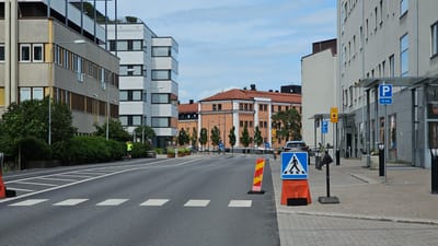 The image depicts a street scene in Linköping, Sweden, likely near the travel center. On the left, a modern grey and beige building with multiple floors and prominent windows lines the street. Adjacent to it, a taller, white building with a distinct architectural style stands. The street itself is paved and marked with pedestrian crossings. A lone individual in a bright green shirt is jogging on the sidewalk on the left side of the road.
In the background, an older, multi-story building with a terracotta-colored roof and white architectural details is visible. This building features numerous windows and appears to be part of an urban complex. Several trees are planted along the sidewalk in front of this building.
On the right side of the street, a grey building with a modern canopy and large windows dominates the foreground. A blue parking sign with a "P" and "10 min" is affixed to a pillar under the canopy. Further down the sidewalk, a yellow sign indicating "Taxi-plats" with "5 min" is visible. A blue accessibility sign is also present.
Several traffic and informational signs are strategically placed along the sidewalk. A prominent blue pedestrian crossing sign, mounted on an orange barrier, is positioned on the right side of the street, facing oncoming traffic. Another striped orange and yellow barrier is located on the road near the pedestrian crossing.
The sky is partly cloudy. The overall impression is one of a well-maintained urban environment with a mix of modern and older architecture, suggesting a functional area within the city.