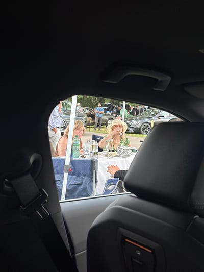 In Ascot, United Kingdom, an outdoor gathering is observed through the interior of a parked vehicle. Two women are seated at a table set with white linen, drinks, and what appears to be food. The woman on the left, with blonde hair, wears a brown sleeveless top and is looking to her right. To her right, another woman, also with blonde hair, is wearing a wide-brimmed straw hat and a green floral dress, and is drinking from a glass. A bottle of water and champagne flutes are visible on the table. Behind them, other attendees and vehicles are present, suggesting an event such as a car show or garden party. The perspective is from inside the vehicle, looking out through the windows, with a portion of the car's interior, including a seatbelt and headrest, framing the scene.