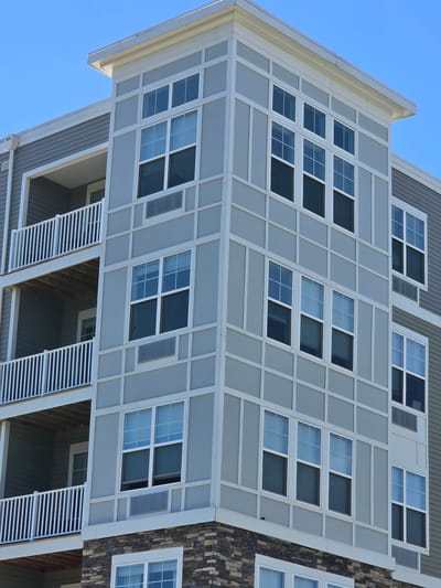 A multi-story apartment building with gray siding and white trim is shown under a clear blue sky. The building features multiple balconies with white railings and several windows. The corner of the building has a decorative paneling design in a lighter gray. The lower portion of the building is clad in stone. The scene is static and does not depict any people or actions. The specific location in Roselle, United States of America is not discernible from the image itself, but the architecture suggests a modern residential complex.