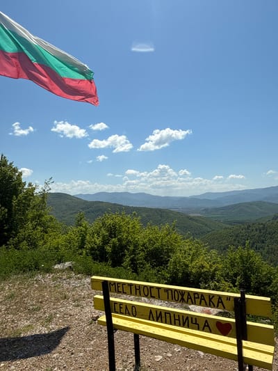 The photograph captures a scenic view from a vantage point in Lipnitsa, Bulgaria. Dominating the foreground is a yellow bench with black lettering that translates to "Place 'Fireplace' Heart Lipnitsa". A red heart is painted between the two lines of text. The background reveals a landscape of rolling, tree-covered hills under a bright blue sky dotted with fluffy white clouds. The Bulgarian flag is visible in the lower left corner. The ground near the bench is covered with gravel. The trees are mostly green.
