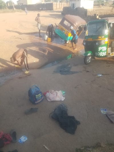 Three shirtless men wash a green and yellow tuk-tuk-style vehicle in an outdoor setting. Water sprays around the vehicle as the men scrub and rinse. The ground appears damp and uneven, with scattered items including clothing, a blue bag, and footwear. Buildings and vegetation are visible in the background under a bright sky.