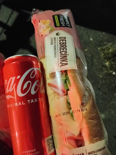 In the city of Popovice, Czechia, a close-up shot reveals a red Coca-Cola can and a packaged sandwich, likely a baguette. The Coca-Cola can is prominently displayed, with its iconic logo visible and the text "ORIGINAL TASTE" and "LAHODNÁ" printed on it. To the right of the can, a sandwich in transparent plastic packaging is shown. The sandwich packaging features text in Czech, including "DEBRECINKA" and "BAGETA S DEBRECINKOU" indicating it contains Debrecin sausage and is a baguette. A date stamp "04.07.2026" is clearly visible on the packaging, suggesting an expiration date. The background is dark and out of focus, but appears to be an outdoor setting with some texture on the ground. The lighting suggests it might be evening or night. No people are visible in the frame.
