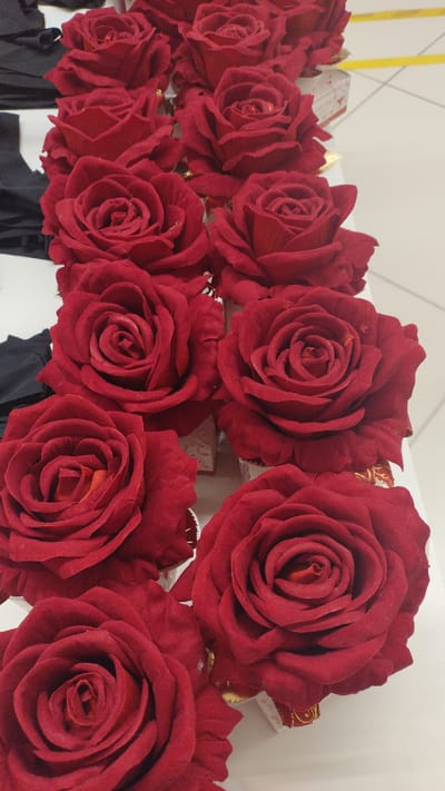 A top-down, eye-level shot shows a display of artificial red roses arranged in multiple rows. The roses are a deep, velvety red with intricate petal formations, and some appear to have gold trim at the base. The background is a white surface, likely a table, with hints of black fabric on the left side. A yellow line can be seen in the distance, suggesting a marked area on a floor or wall. The setting appears to be indoors, possibly a retail or display area. The roses are the primary focus, presented in a visually rich and orderly fashion.