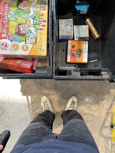 This top-down image depicts a person standing on a concrete floor in the Polígon Industrial De Son Castello, Spain, looking down into two open, black plastic storage crates positioned directly in front of them. The person is wearing a dark blue shirt, dark grey wide-leg pants, and off-white sneakers with a star emblem. They are holding a black-cased smartphone in their left hand. The concrete floor shows wear, cracks, and some irregular discoloration, with a yellow marking line visible on the right.
The left crate contains a large, colorful box for a "Kit de Ciencias" (Science Kit) with cartoon illustrations of children, positioned atop a smaller red rectangular box. The right crate contains an assortment of items, including an HP 303 ink cartridge, a gold-colored aerosol can of L'Oreal hair product, several small packaged items (likely electronic components), and an orange and black box labeled "GR.FEMA Bike Cable Lock." A small white slip of paper is also visible within the right crate. Strong overhead lighting casts shadows of the crates and the person onto the floor.