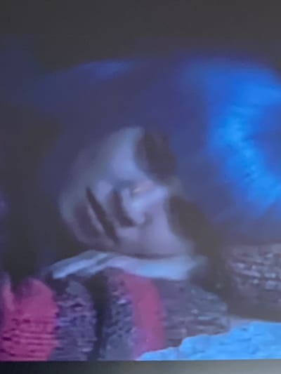 A close-up shot shows a person with bright blue hair, likely a wig or dyed hair, looking down with their eyes partially closed. The person's face is partially obscured by their hair and a textured, possibly knitted, object in the foreground, which appears to be a piece of clothing, perhaps a scarf or a glove, with stripes of red, pink, and darker purple or black. The lighting is dim, with a strong blue hue cast across the scene, suggesting artificial lighting or a specific filter. The overall mood is somewhat somber or introspective, given the subject's posture and the subdued lighting. There are no visible text elements in the image. The context provided places this image in Baošići, Montenegro, though there are no direct visual cues in the image to confirm this specific location.