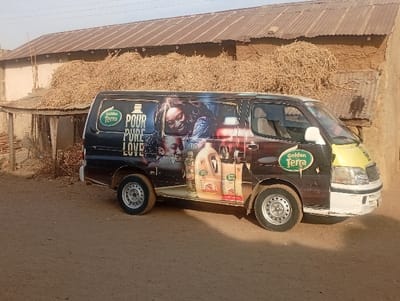 A van, painted with a “Golden Terra” advertisement featuring a woman and child, is parked outdoors. The backdrop includes an old building with a corrugated metal roof partially covered with straw or hay. The vehicle's front is predominantly white and yellow, contrasting with the dark body adorned with product images. The ground is dusty, and a glimpse of a rudimentary shed is visible on the left.